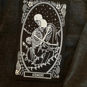 May include: Black and white embroidered patch depicting two skeletons embracing in a starry night sky. The patch is framed with a decorative border and the word "GEMINI" is printed at the bottom.