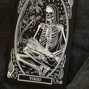 May include: Black and white embroidered patch depicting a skeleton holding wheat stalks. The patch is surrounded by a floral design and the word "VIRGO" is printed at the bottom.