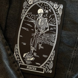 May include: A black and white embroidered patch depicting a skeleton sitting on a pier, with the word "Pisces" at the bottom. The patch is surrounded by a floral design.
