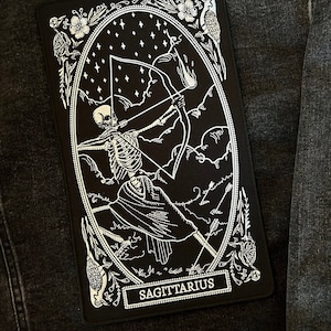May include: Black and white embroidered iron-on patch featuring a skeletal figure with an arrow and bow, set against a starry night sky. The patch is framed with floral details and the text "SAGITTARIUS" at the bottom.