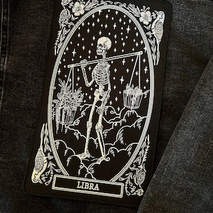 May include: Black and white embroidered patch featuring a skeleton holding a scale with the word "LIBRA" at the bottom.