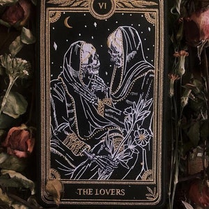 May include: A black and gold embroidered tarot card depicting the Lovers card, featuring two skeletons in hooded robes holding a flower. The card number VI is in the top right corner.