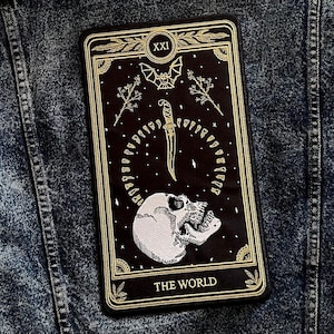May include: A black and gold embroidered patch depicting the World tarot card. The card features a skull, a dagger, a bat, and the text "XXI THE WORLD".