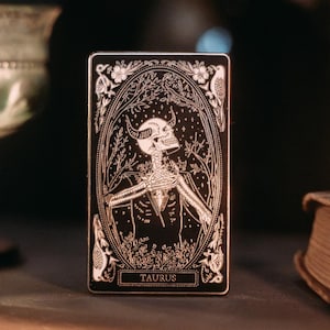 May include: A black tarot card with a silver design depicting a skeleton with horns and the word "Taurus" at the bottom.