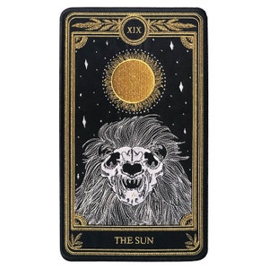 May include: A black and gold tarot card with the number XIX and the words "THE SUN" printed on it. The card features a white embroidered image of a lion's skull with a mane of hair.