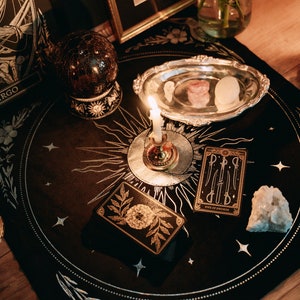 May include: A black velvet tarot cloth with gold embroidery of a sun, moon, and stars. A lit candle sits in a silver holder on the cloth. There are tarot cards, a crystal, and a silver dish with crystals on the cloth. The text on one of the cards reads "Five of Swords".