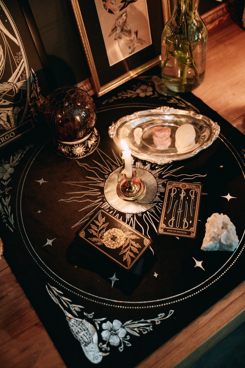 May include: A black velvet tarot cloth with gold embroidery of a sun, moon, and stars. A lit candle sits in a silver holder on the cloth. There are tarot cards, a crystal, and a silver dish with crystals on the cloth. The text on one of the cards reads "Five of Swords".