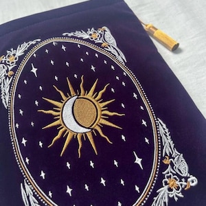 May include: A dark blue velvet pouch with an embroidered celestial design. The design features a gold sun and crescent moon, surrounded by stars and floral accents. A gold tassel hangs from the top. The pouch is likely used for storing small items.