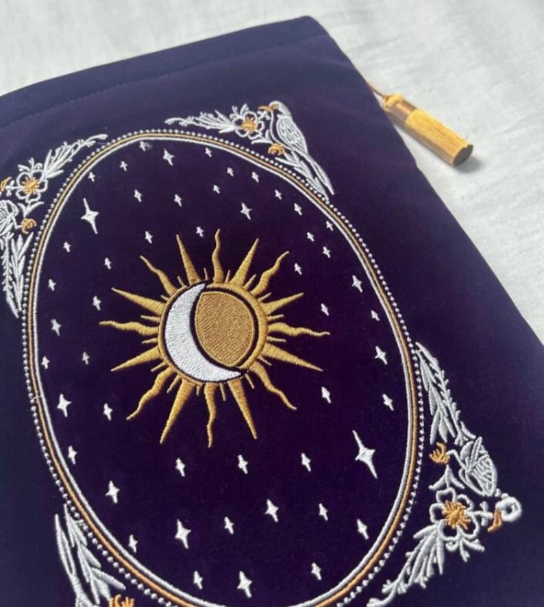 May include: A dark blue velvet pouch with an embroidered celestial design. The design features a gold sun and crescent moon, surrounded by stars and floral accents. A gold tassel hangs from the top. The pouch is likely used for storing small items.