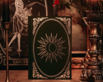 May include: A dark green hardcover book with gold accents and a sun and moon design on the front cover. The book is sitting on a wooden table with candles and other decorative items.