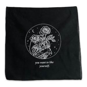 May include: Black bandana with a white line drawing of a skull with roses in its mouth. The skull is inside a circle with a crescent moon and stars. The text "you want to like yourself." is below the circle.