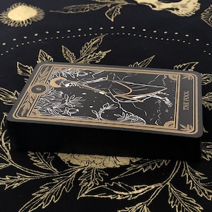 May include: A deck of tarot cards with a black background and gold accents. The card on top features an illustration of a figure with the words "THE FOOL". The deck is resting on a black cloth with a gold floral pattern.