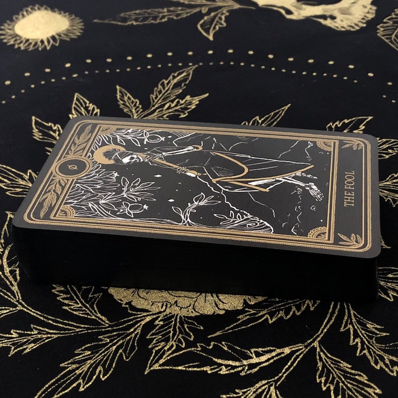 May include: A deck of tarot cards with a black background and gold accents. The card on top features an illustration of a figure with the words "THE FOOL". The deck is resting on a black cloth with a gold floral pattern.