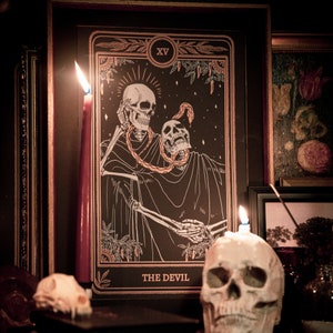 Puede incluir: Una carta de tarot vintage con una ilustración en blanco y negro de dos esqueletos con una serpiente enroscada alrededor de ellos. La carta se titula "El Diablo" y tiene el número XV en la esquina superior derecha.