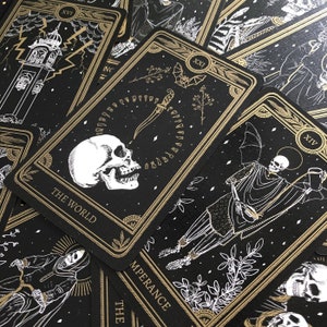 May include: A collection of tarot cards with black backgrounds and gold accents. The cards feature illustrations of skulls, skeletons, and other gothic imagery. The card labeled "The World" is prominently displayed, along with other cards with Roman numerals.