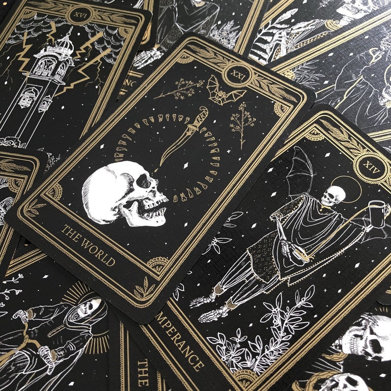May include: A collection of tarot cards with black backgrounds and gold accents. The cards feature illustrations of skulls, skeletons, and other gothic imagery. The card labeled "The World" is prominently displayed, along with other cards with Roman numerals.