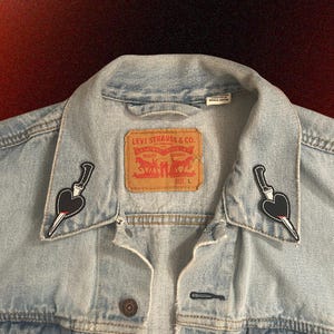 May include: Light blue denim jacket with two black and white patches of a knife piercing a heart on the collar. The jacket has a Levi Strauss & Co. label and a red tag. Size L.