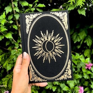 May include: Black journal with a gold sun and moon design on the cover. The design is surrounded by a gold ornate frame.