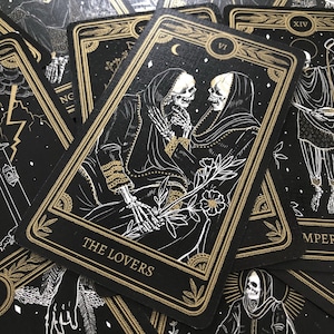 May include: A collection of tarot cards with a black background and gold accents. The central card, labeled "The Lovers," features skeletal figures in cloaks, with a floral element. Other cards are visible, each with unique illustrations and Roman numerals.