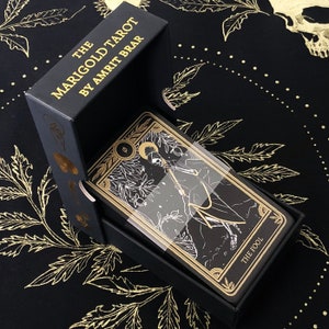 May include: A black tarot card deck titled "The Marigold Tarot by Amrit Brar" in an open black box. The cards feature gold and white illustrations on a black background. The card on top is labeled "THE FOOL". The box and cards are on a black cloth with gold leaf designs.