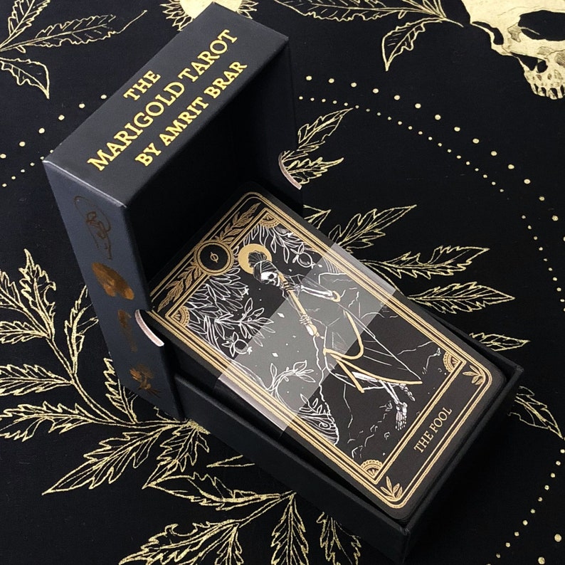 May include: A black tarot card deck titled "The Marigold Tarot by Amrit Brar" in an open black box. The cards feature gold and white illustrations on a black background. The card on top is labeled "THE FOOL". The box and cards are on a black cloth with gold leaf designs.