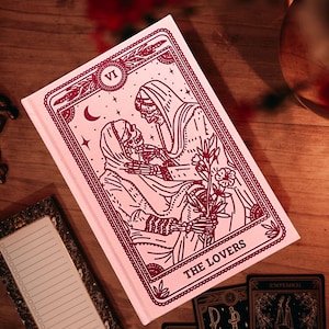May include: A pink journal with a red illustration of two skeletal figures embracing, holding a flower, and the words "THE LOVERS". The journal is surrounded by tarot cards and a notepad on a wooden surface. The journal has the Roman numeral "VI" at the top.