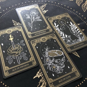 May include: Four tarot cards with gold and white illustrations on a black background. The cards are labeled "Ace of Wands," "Ace of Swords," "Ace of Rings," and "Ace of Cups." The cards feature floral and skull imagery.