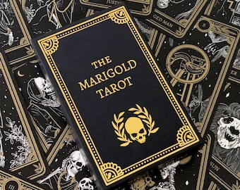 以下が含まれることがあります： 金色の縁取りと「THE MARIGOLD TAROT」の文字が金色で書かれた黒いタロットカードの箱。金色のドクロと月桂樹のデザインがテキストの下にあります。箱は、金と白のイラストが描かれたタロットカードに囲まれています。