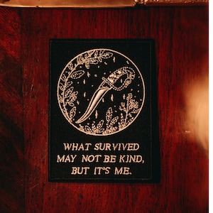 May include: Black embroidered patch with a dagger design inside a circle of leaves and stars. The text reads "WHAT SURVIVED MAY NOT BE KIND, BUT IT'S ME." The patch is displayed on a dark wooden surface.