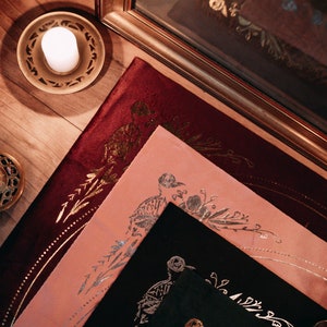 May include: Three decorative fabric squares with gold and silver metallic designs. The squares are layered on top of each other, with the top square being black, the middle square being pink, and the bottom square being burgundy.