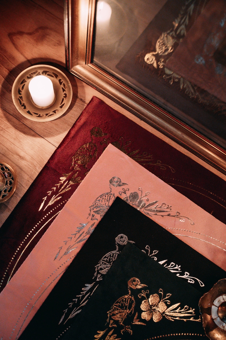 May include: Three decorative fabric squares with gold and silver metallic designs. The squares are layered on top of each other, with the top square being black, the middle square being pink, and the bottom square being burgundy.