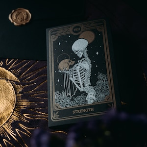 May include: A black tarot card with gold accents depicting a skeleton holding a skull. The card is labeled "VIII STRENGTH".