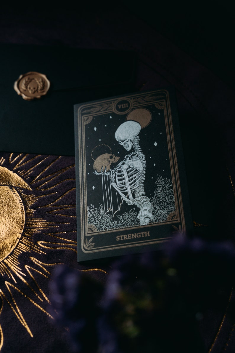 May include: A black tarot card with gold accents depicting a skeleton holding a skull. The card is labeled "VIII STRENGTH".