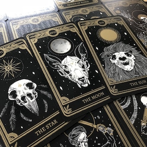 May include: A deck of tarot cards with black backgrounds and gold accents. The cards feature illustrations of skulls, celestial bodies, and botanical elements. The cards are labeled with titles such as "The Moon" and "The Star".
