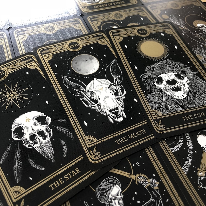 May include: A deck of tarot cards with black backgrounds and gold accents. The cards feature illustrations of skulls, celestial bodies, and botanical elements. The cards are labeled with titles such as "The Moon" and "The Star".