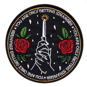 Peut inclure: Patch brodé noir avec une main blanche tenant un couteau avec une flamme au bout. Le patch a deux roses rouges avec des feuilles vertes de chaque côté de la main. Le texte autour du bord du patch est "You are only getting stranger + You are only getting stranger".