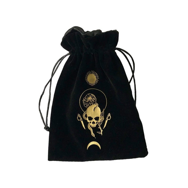 May include: Black velvet pouch with a gold skull and snake design. The pouch features a sun, moon, and floral accents.