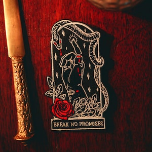 May include: A black embroidered patch with a hand, snake, and rose design. The hand has a cut finger, and the snake is wrapped around the frame. The text "BREAK NO PROMISES" is at the bottom. The patch is on a dark wood surface.