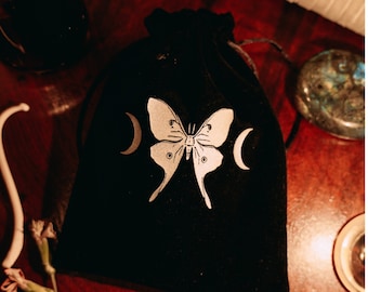 May include: Black velvet pouch with a silver moon and butterfly design. The pouch is closed with a drawstring.