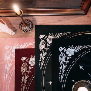 May include: Three fabric placemats with a black, burgundy, and pink background. Each placemat features a silver metallic design of a bird with flowers and leaves. The design is repeated on each placemat.
