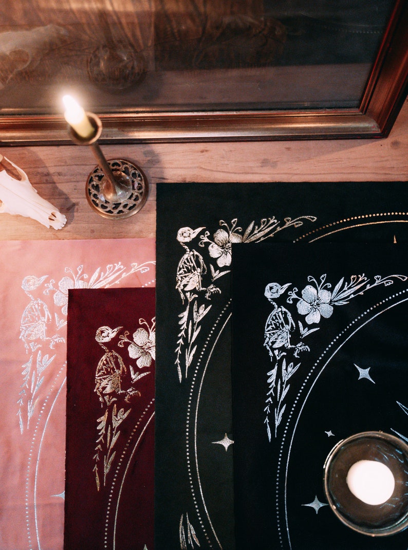May include: Three fabric placemats with a black, burgundy, and pink background. Each placemat features a silver metallic design of a bird with flowers and leaves. The design is repeated on each placemat.