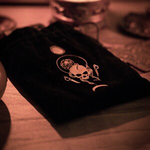 May include: A black velvet pouch with a white skull and crescent moon design. The pouch is on a wooden surface.