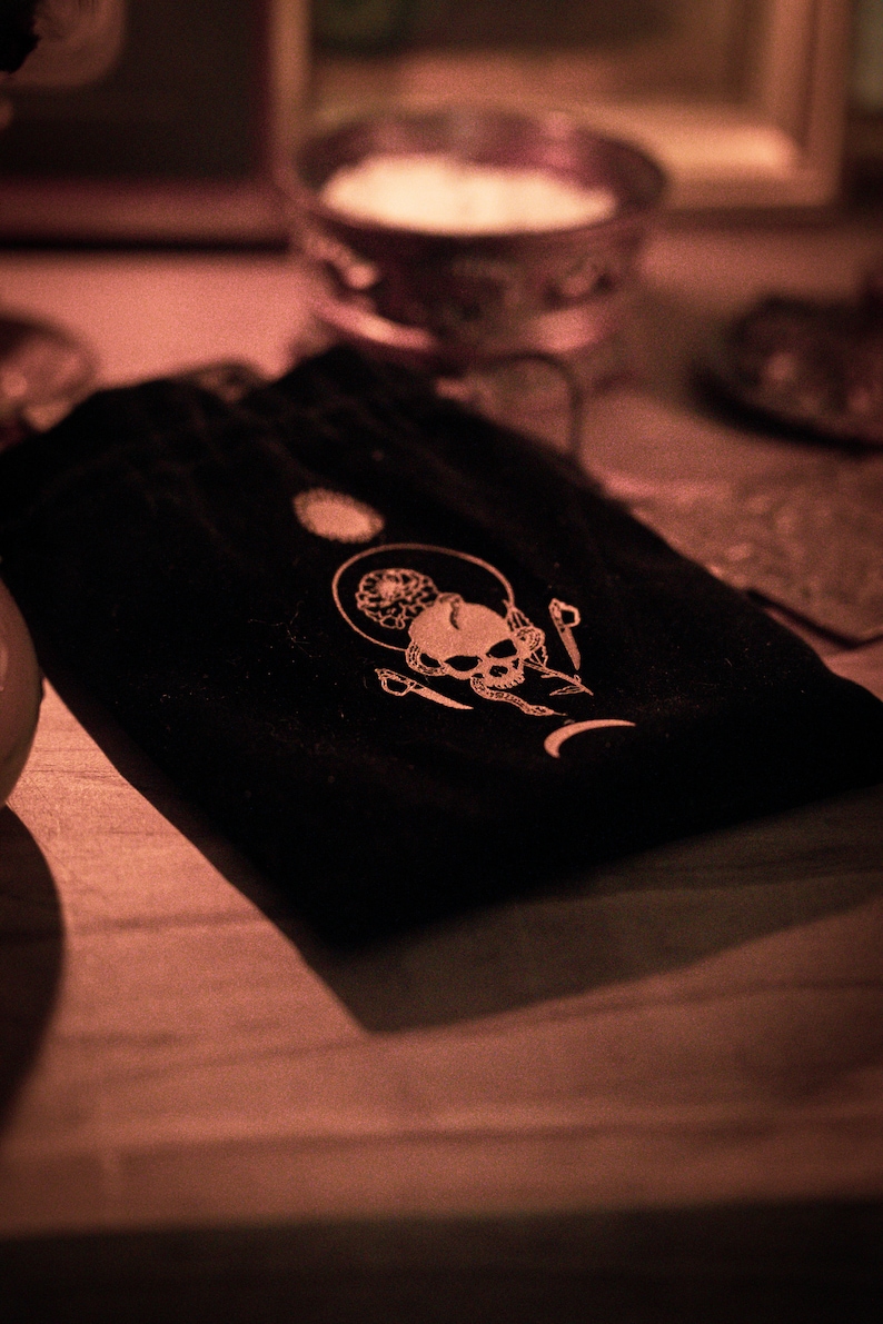 May include: A black velvet pouch with a white skull and crescent moon design. The pouch is on a wooden surface.