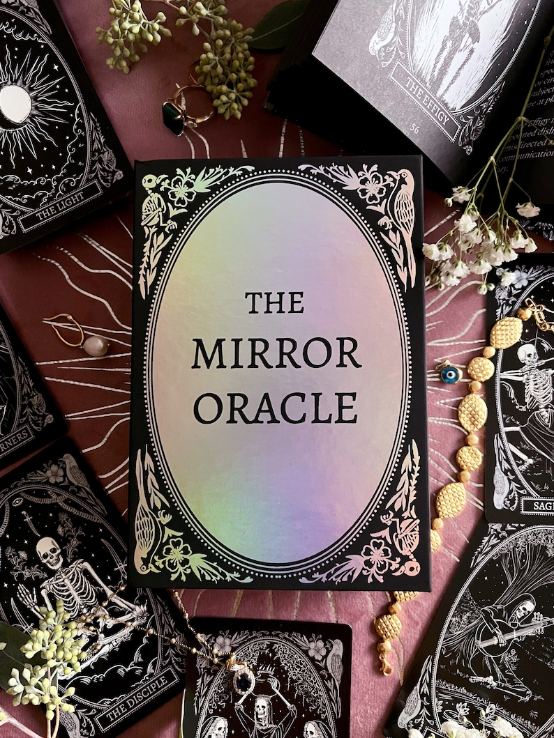 Peut inclure: Une bo&icirc;te noire avec un ovale holographique iridescent au centre. La bo&icirc;te est orn&eacute;e d'un motif noir et blanc avec des motifs floraux et d'oiseaux. Le texte "THE MIRROR ORACLE" est imprim&eacute; en blanc sur la bo&icirc;te.