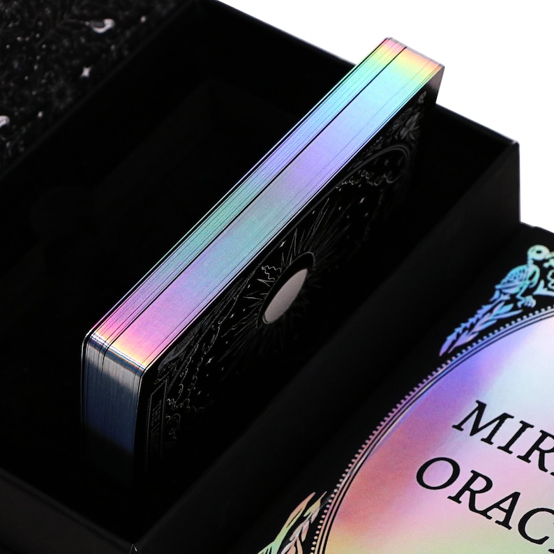 Peut inclure: Un jeu de cartes de tarot noir et argent avec une finition holographique iris&eacute;e. Les cartes sont dans une bo&icirc;te noire avec les mots "Mirror Oracle" imprim&eacute;s sur le devant.