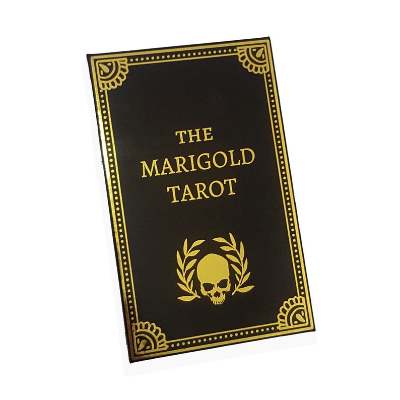 May include: A black tarot card deck titled "The Marigold Tarot." The cover features gold lettering and decorative gold accents. A gold skull and laurel wreath design is centered at the bottom.