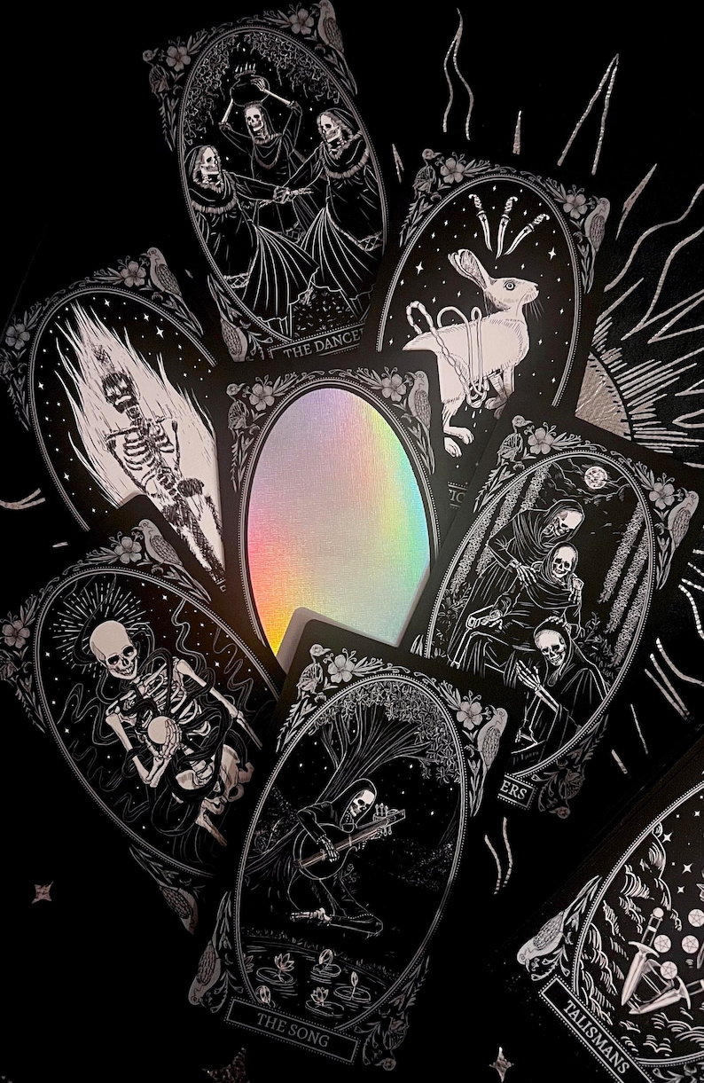 Peut inclure: Un jeu de cartes de tarot noires avec des accents argent&eacute;s et des illustrations de squelettes, un lapin et une figure &agrave; capuchon. Les cartes portent des titres tels que "La Danse", "Le Chant", "Talisman" et "Feux".
