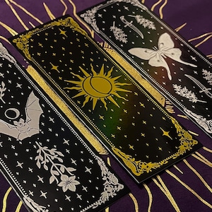 May include: Three black and gold bookmarks with celestial and botanical designs. One bookmark features a bat, another a sun, and the third a moth. Each bookmark has a gold border and white accents, with a starry background.
