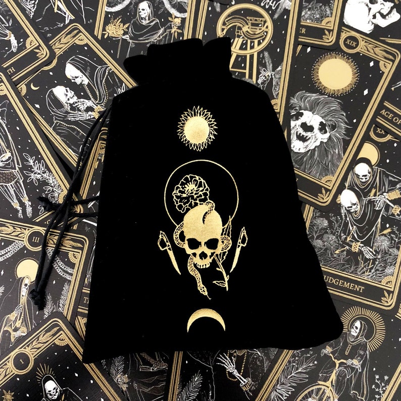 May include: Black velvet bag with a gold skull and snake design. The bag is surrounded by tarot cards with gold accents and black and white illustrations.