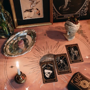 May include: A tarot card reading spread with four cards laid out on a pink table cloth with a silver sun and moon design. The cards are "Knight of Cups", "Queen of Swords", "Page of Wands", and "Page of Death".